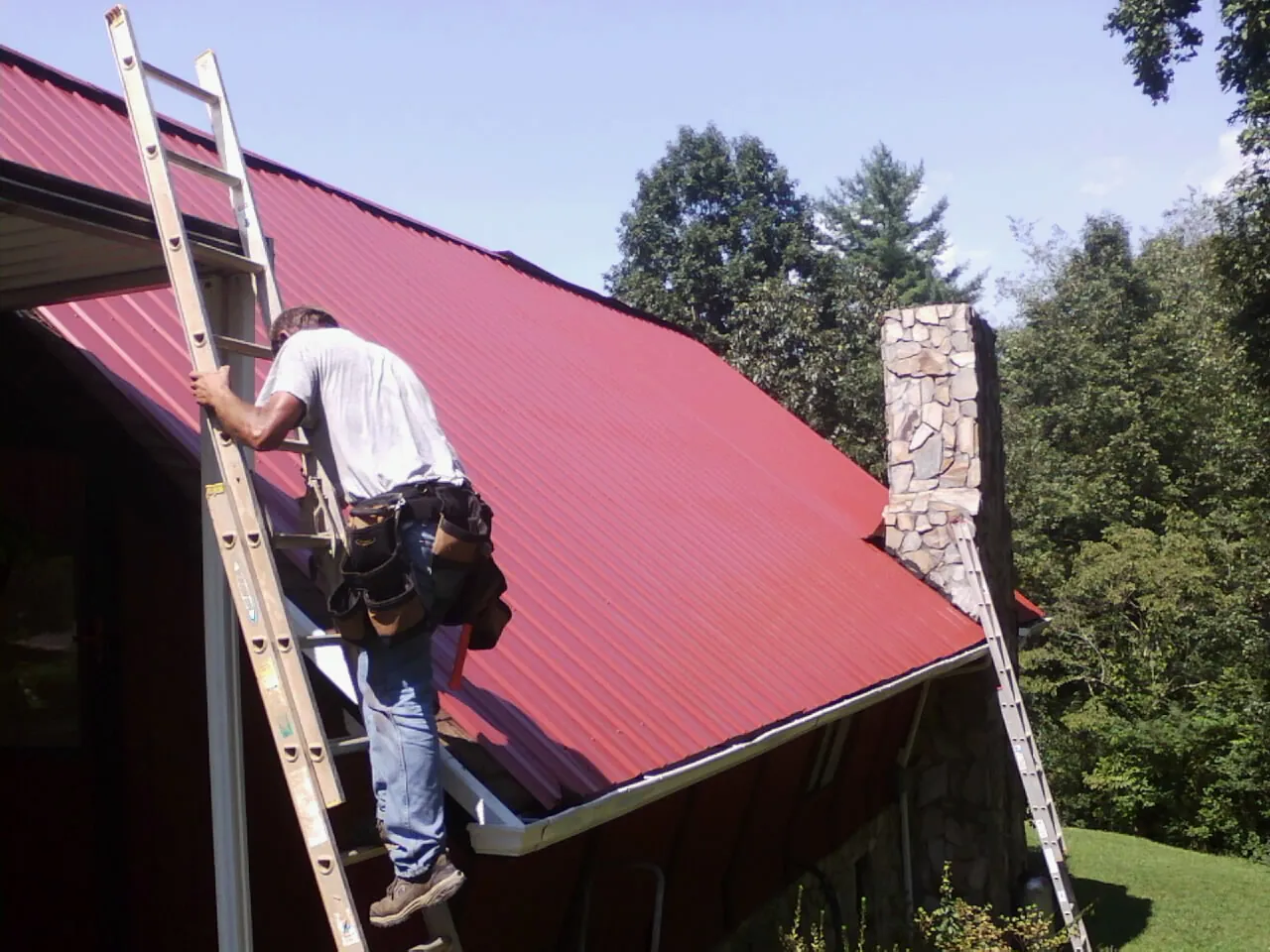 Roofer on a ladder beside a red metal roof and stone chimney — Soffit & Fascia Repair by Roofing Brothers in Sassamansville